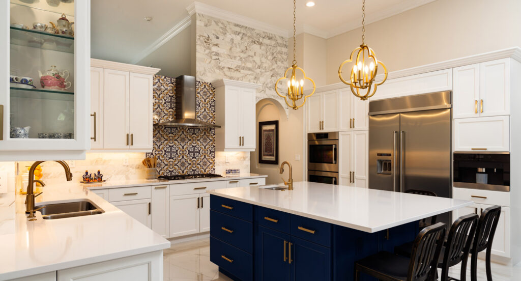 Modern kitchen with white cabinets, blue island, stainless steel appliances, gold fixtures, patterned backsplash, and pendant lighting.