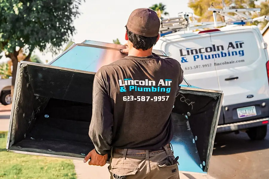 A worker in a "Lincoln Air & Plumbing" shirt carries a large metal duct toward a company van parked on a residential street.