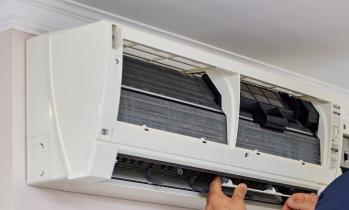 A person is servicing an open wall-mounted air conditioning unit, with the front panel lifted to access the internal components.