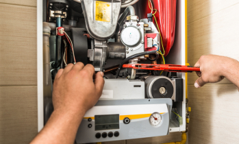 A person uses a wrench to repair or adjust components inside an open gas boiler unit mounted on a wall.