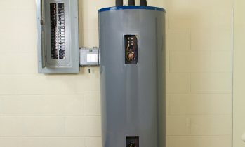 A gray electric water heater stands next to an electrical breaker panel mounted on a beige wall.
